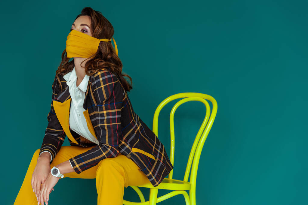 модна жінка в жовтій масці і плетений блейзер позує, сидячи на стільці ізольовані на синьому
 - Фото, зображення