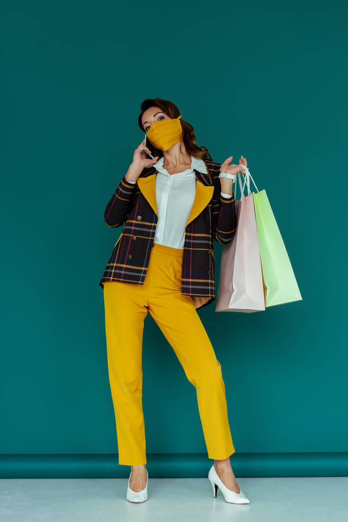 elegante donna in maschera e blazer a quadri in posa con borse della spesa e parlando su smartphone su blu
 - Foto, immagini