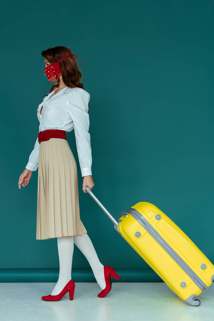κομψή νεαρή γυναίκα σε μάσκα με τα πόδια με ταξιδιωτική τσάντα σε μπλε  - Φωτογραφία, εικόνα