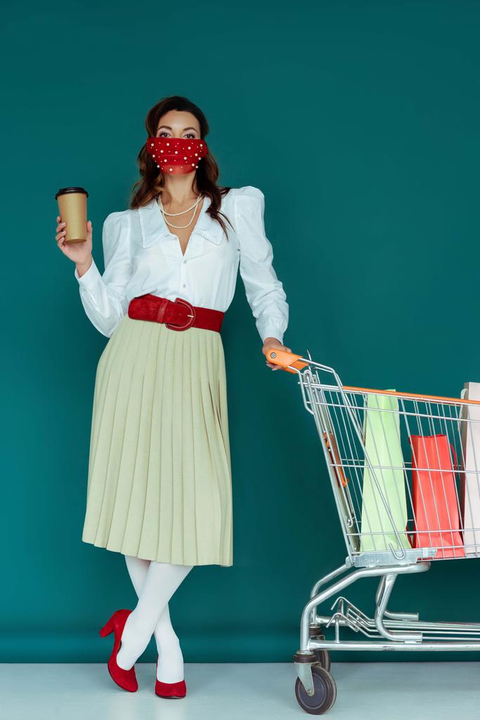chica con estilo en la máscara de la celebración de café para ir y de pie cerca del carrito de la compra con bolsas de compras en azul
  - Foto, Imagen