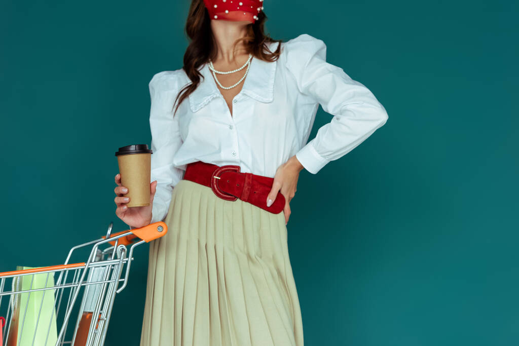 przycięty widok modnej dziewczyny w czerwonej masce trzymając kawę iść i stojąc z ręką na biodrze w pobliżu koszyka zakupów z torby na zakupy odizolowane na niebiesko  - Zdjęcie, obraz