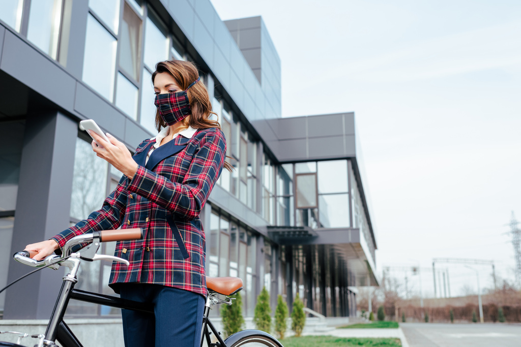 mujer de negocios en máscara de cuadros con teléfono inteligente cerca de la bicicleta fuera
  - Foto, Imagen