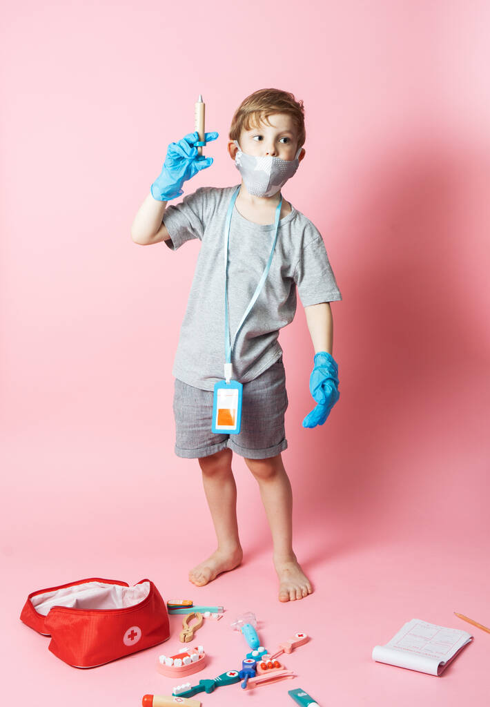 Środki ochrony indywidualnej: mały chłopiec w masce medycznej i niebieskie rękawiczki bawi się zestawem dziecięcym lekarza. - Zdjęcie, obraz