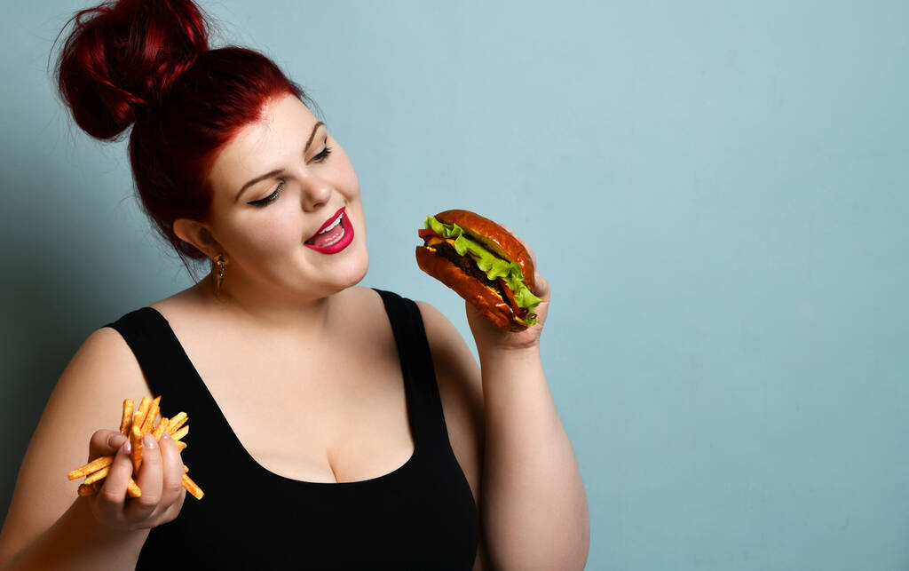 Feliz excesso de peso mulher gorda feliz segurar hambúrguer cheeseburger sanduíche com carne bovina e batatas fritas na mão
 - Foto, Imagem