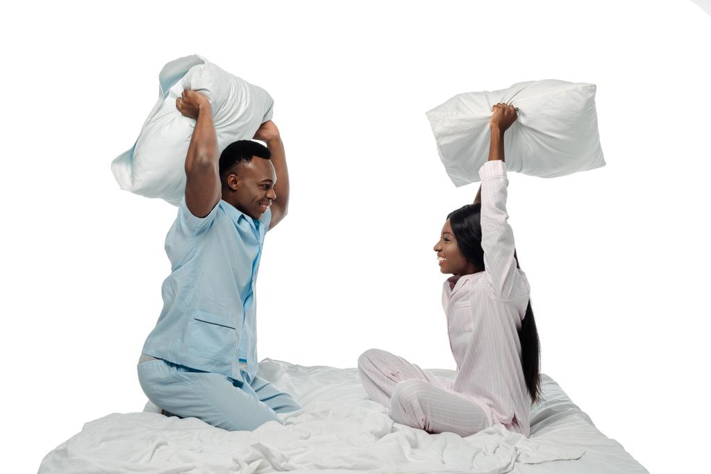 feliz africano americano pareja teniendo almohada lucha en cama en pijama aislado en blanco
 - Foto, Imagen