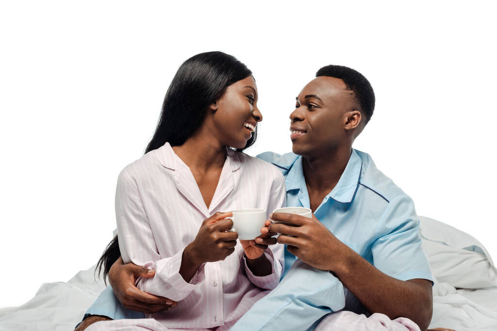 Mutlu Afro-Amerikan çifti beyaz pijamalarıyla yatakta kahve içiyorlar. - Fotoğraf, Görsel