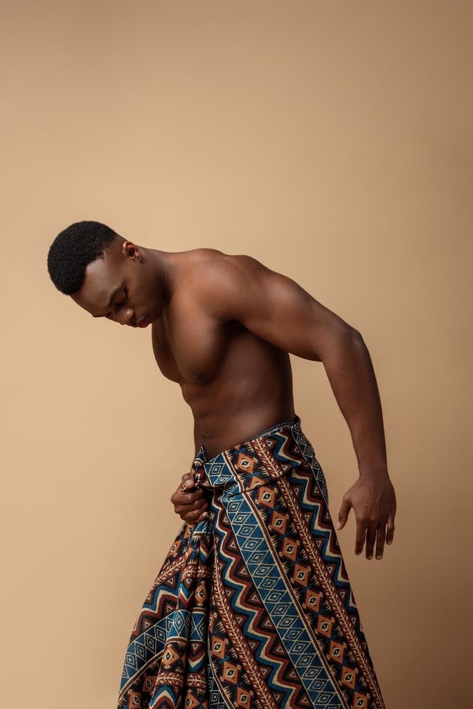 сексуальний голий племінний афро-чоловік вкритий ковдрою позує на бежевому
 - Фото, зображення