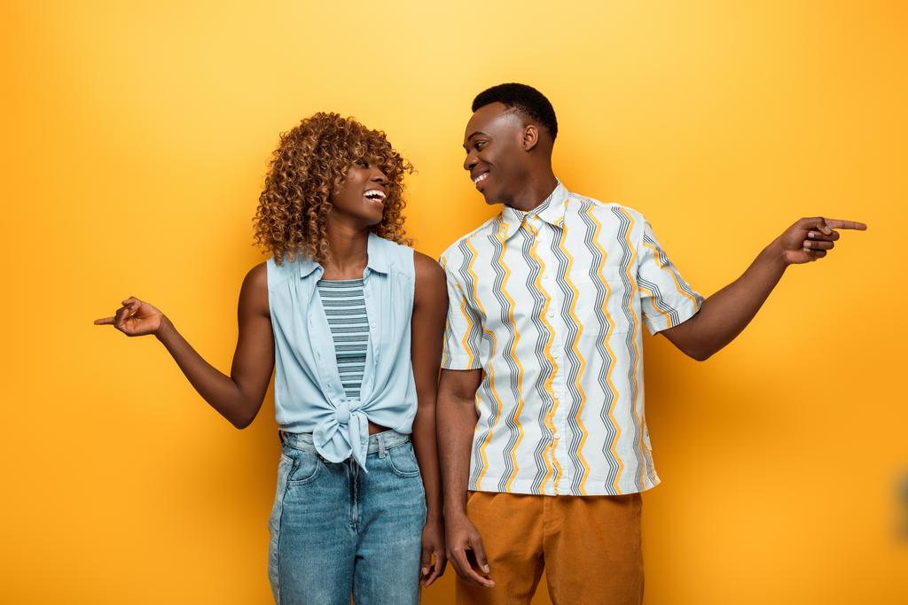 šťastný africký americký pár ukazuje prsty a dívá se na sebe na žluté barevné pozadí - Fotografie, Obrázek