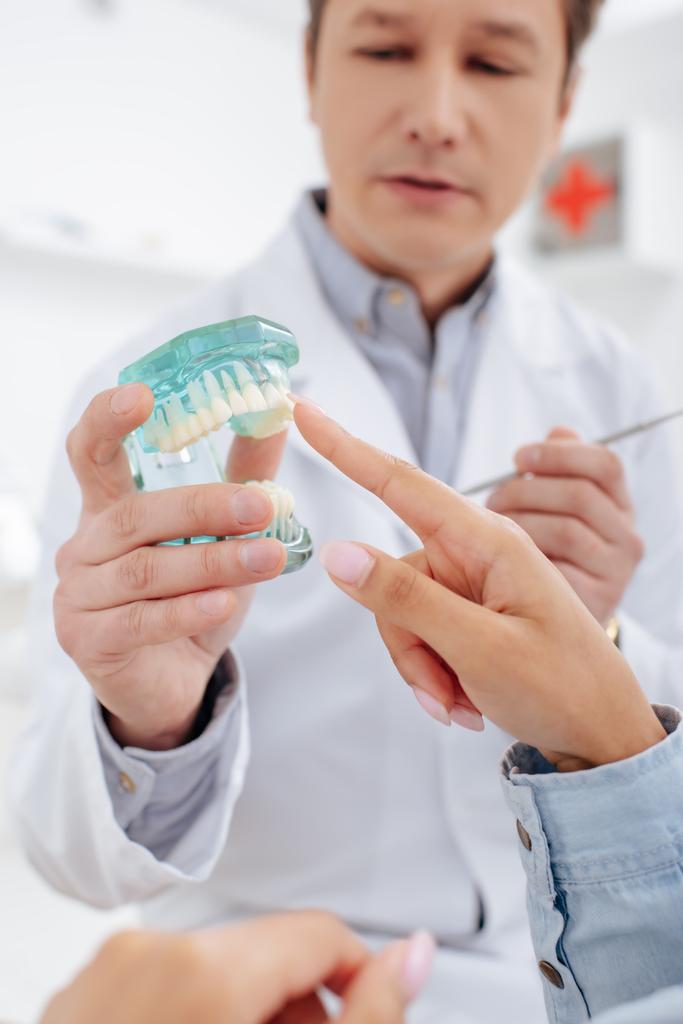 Selektiver Fokus des Zahnarztes im weißen Kittel in der Nähe des Patienten, der mit dem Finger auf das Zahnmodell zeigt  - Foto, Bild
