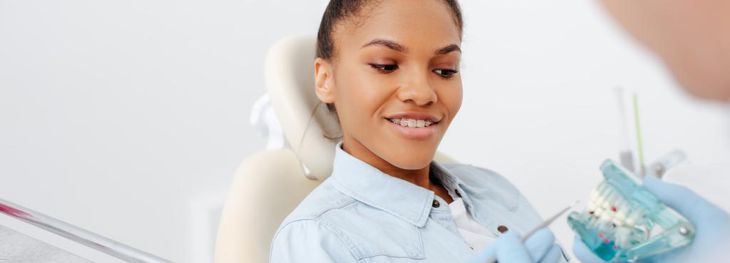 tiro panorâmico de mulher americana africana feliz em aparelho perto dentista segurando dentes modelo
  - Foto, Imagem