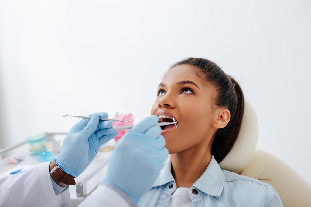 Zahnarzt in Latexhandschuhen mit Zahnausrüstung in der Nähe eines afrikanisch-amerikanischen Patienten mit geöffnetem Mund und Zahnspange  - Foto, Bild