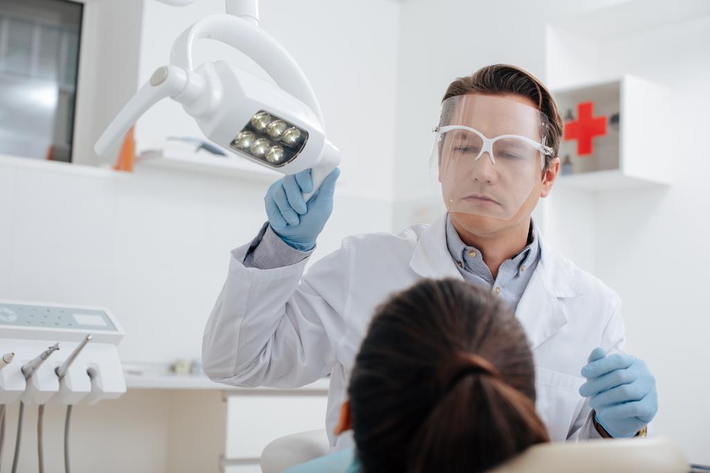 вибірковий фокус стоматолога на щиті та латексних рукавичках, що торкаються медичної лампи біля жінки
  - Фото, зображення