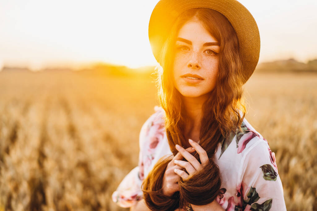 Portré egy gyönyörű fiatal nőről göndör hajjal és szeplős arccal. Nő ruha és kalap pózol búzamezőn naplementekor és a kamerába néz. - Fotó, kép