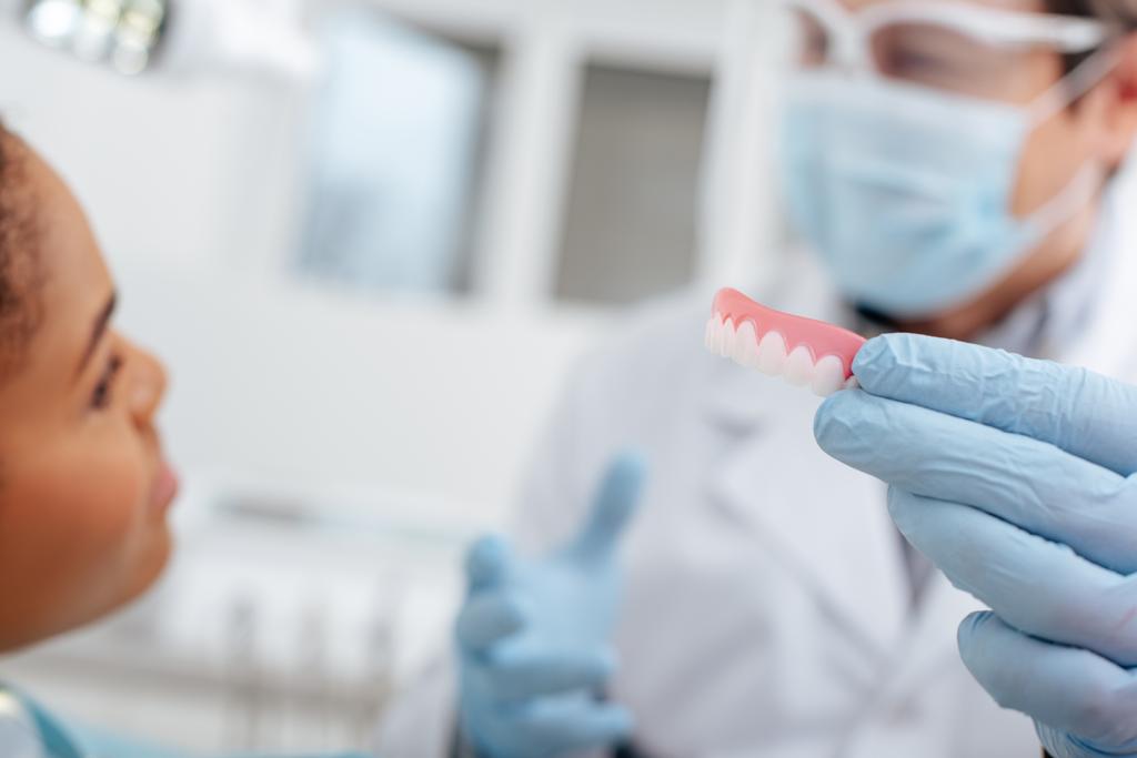 селективный фокус стоматолога в латексных перчатках, держащих зубной протез рядом с африканской американкой
  - Фото, изображение
