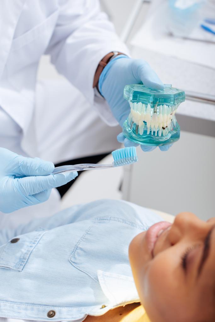 selektywne skupienie się dentysty trzymającego model zębów i szczoteczkę do zębów w pobliżu szczęśliwego amerykańskiego pacjenta  - Zdjęcie, obraz