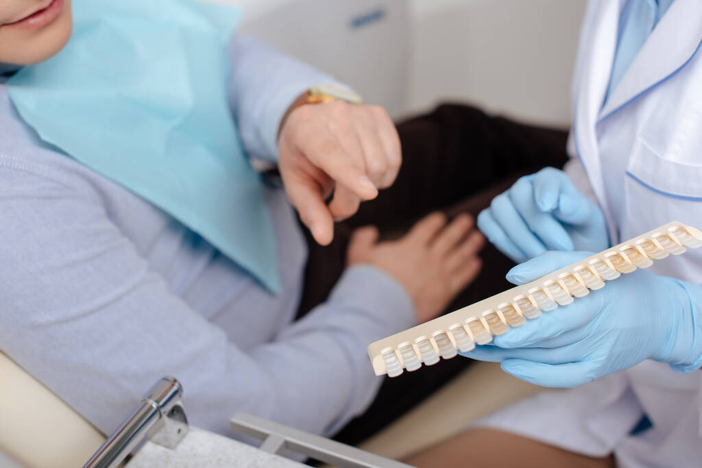 обрезанный вид человека, указывающего пальцем на палитру зубов возле стоматолога
  - Фото, изображение