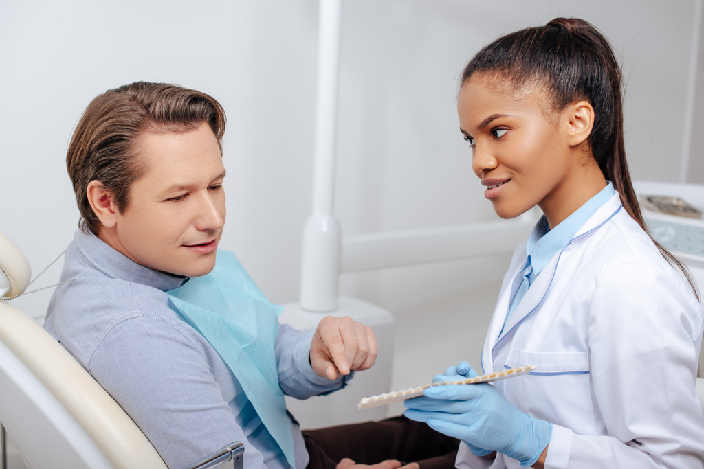 hombre apuntando con el dedo a la paleta de dientes cerca del feliz dentista afroamericano en guantes de látex
 - Foto, Imagen