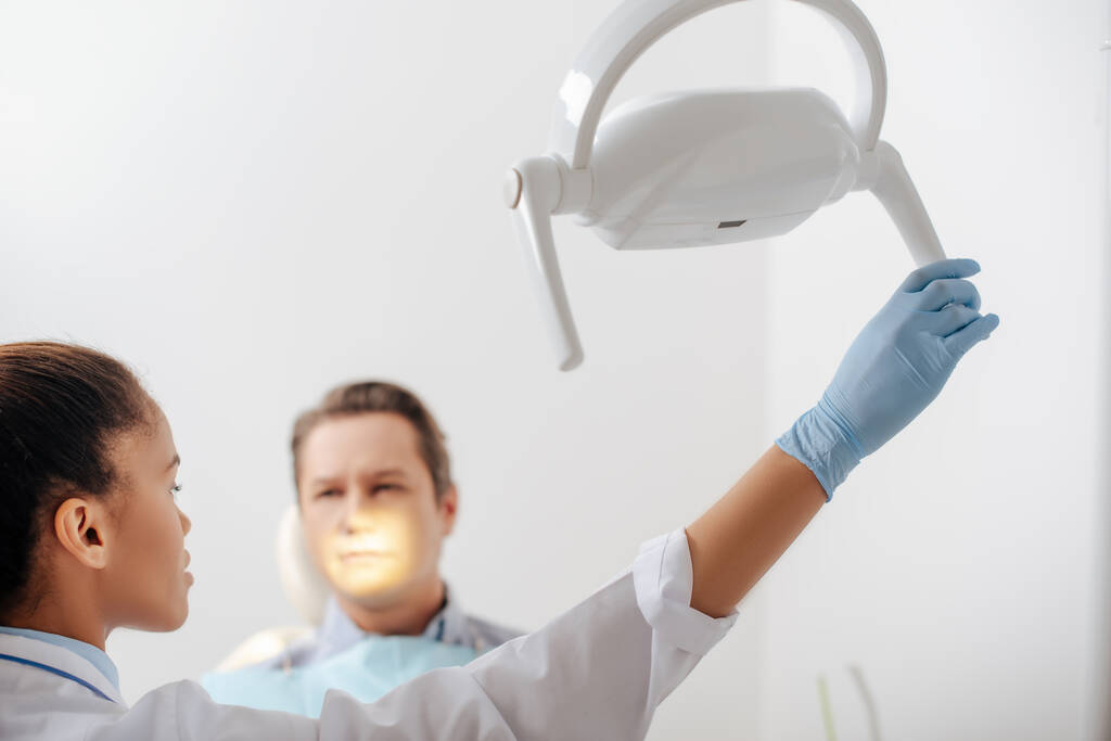 επιλεκτική εστίαση των ελκυστικών αφροαμερικανός οδοντίατρος στο γάντι λατέξ αγγίζοντας ιατρική λάμπα κοντά στον ασθενή  - Φωτογραφία, εικόνα