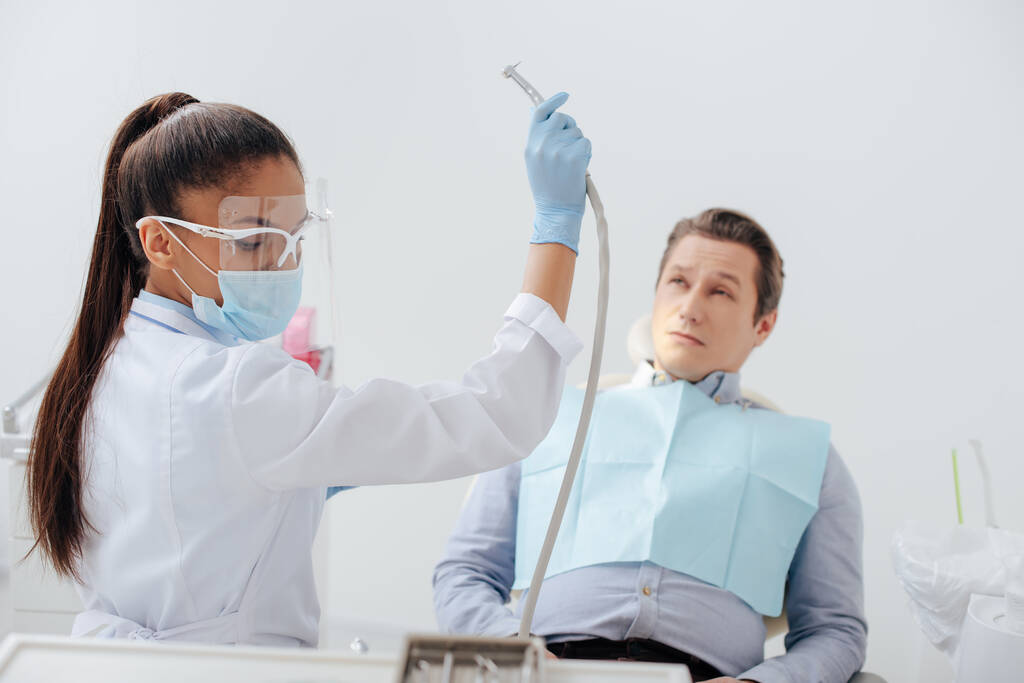 Африканский американский стоматолог в медицинской маске и щит для лица держит стоматологический инструмент рядом с человеком
  - Фото, изображение