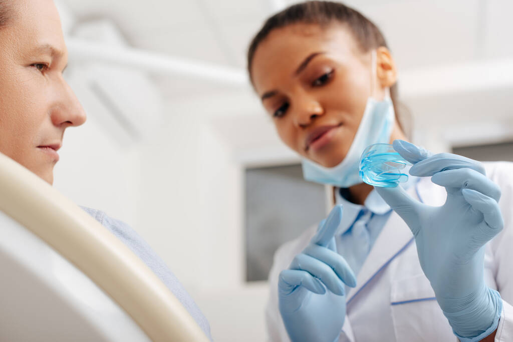 wybiórcze skupienie afrykańskiego amerykańskiego dentysty w rękawiczkach lateksowych wskazujących palcem na aparat w pobliżu pacjenta  - Zdjęcie, obraz