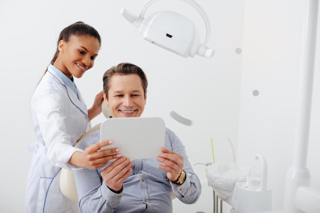 paciente feliz olhando para o espelho perto alegre dentista afro-americano em aparelho
 - Foto, Imagem