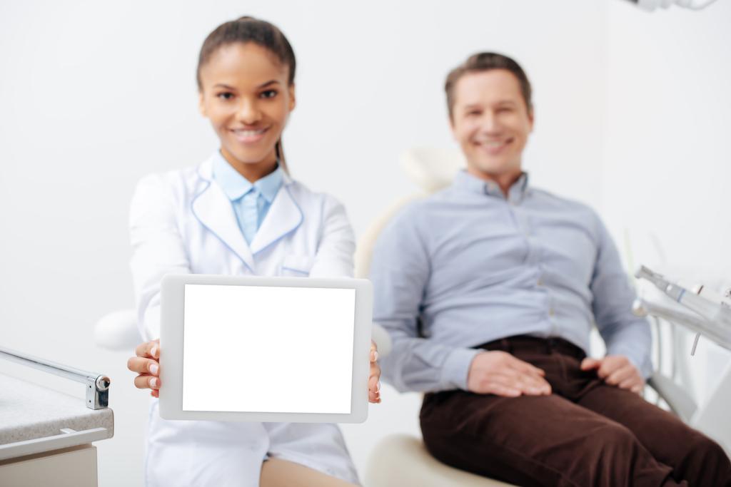 selective focus of smiling african american dentist holding digital tablet with white screen near happy patient  - Photo, Image