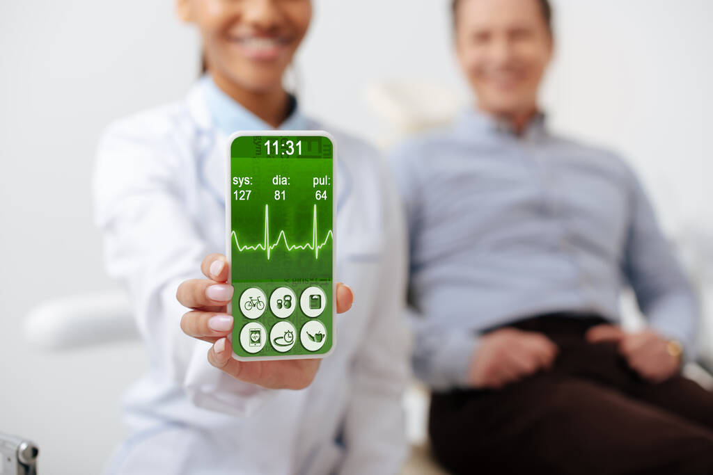 foyer sélectif de joyeux dentiste afro-américain tenant smartphone avec rythme cardiaque à l'écran près patient heureux
  - Photo, image