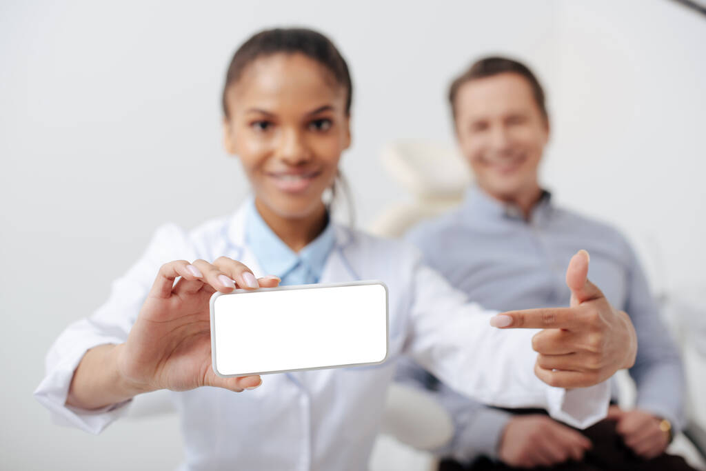 messa a fuoco selettiva di allegro dentista afroamericano che punta con il dito allo smartphone con schermo bianco vicino al paziente felice
  - Foto, immagini