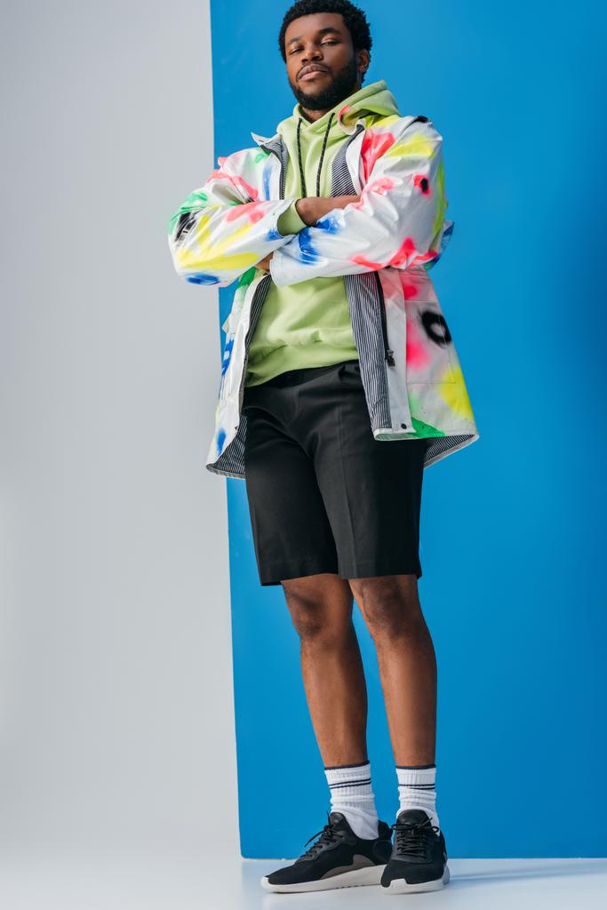 modny Afrykański Amerykanin pozowanie z skrzyżowanymi ramionami w kolorowy futurystyczny wygląd na szary i niebieski - Zdjęcie, obraz