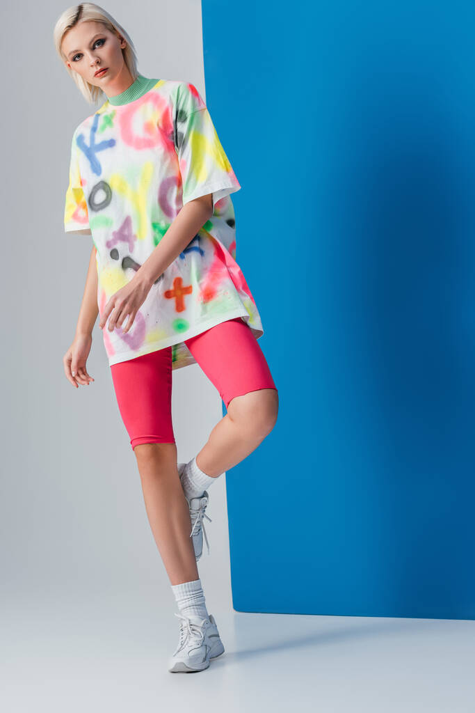 ragazza bionda alla moda in posa in pantaloncini da bici rosa fluo e t-shirt colorata su grigio e blu
  - Foto, immagini