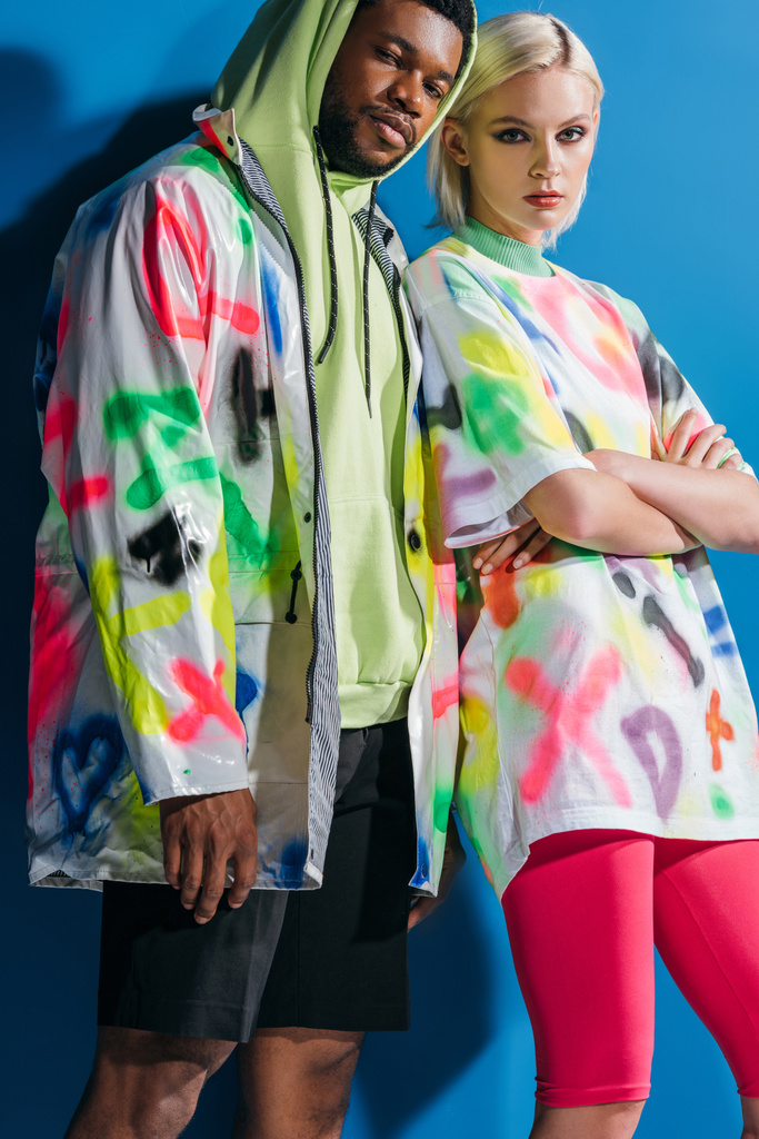 стильная межрасовая пара в красочном футуристическом виде позирует со скрещенными руками на голубом
 - Фото, изображение