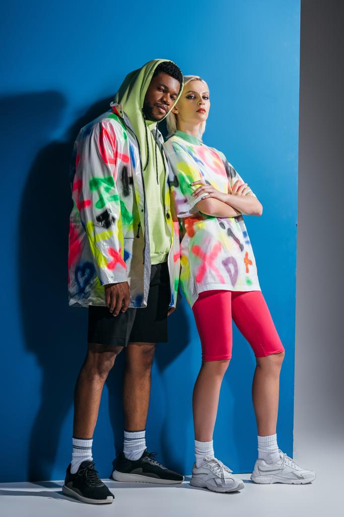 modisches multikulturelles Paar posiert in buntem futuristischen Look auf Grau und Blau - Foto, Bild