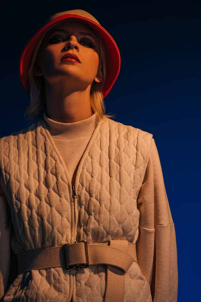 modny model pozowanie w futurystyczny wygląd i kapelusz na granatowym - Zdjęcie, obraz