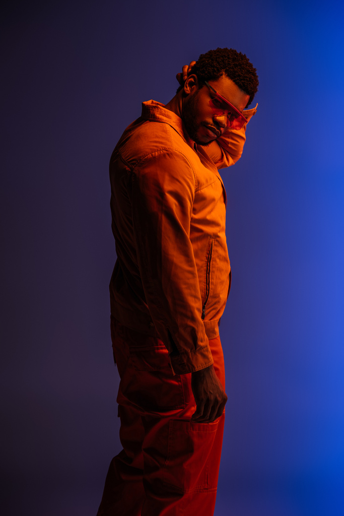 uomo afroamericano in look futuristico e occhiali da sole in posa su blu a luce rossa
  - Foto, immagini