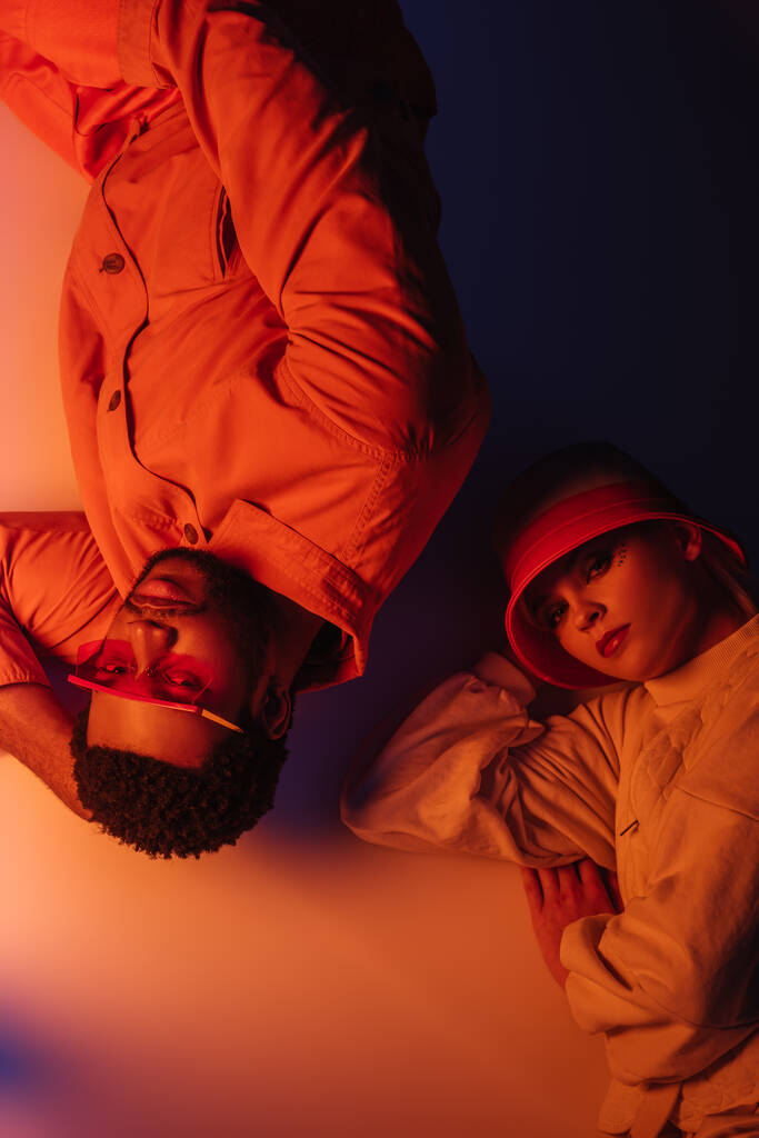 vue de dessus du couple interracial à la mode en vêtements futuristes couché dans la lumière orange
 - Photo, image