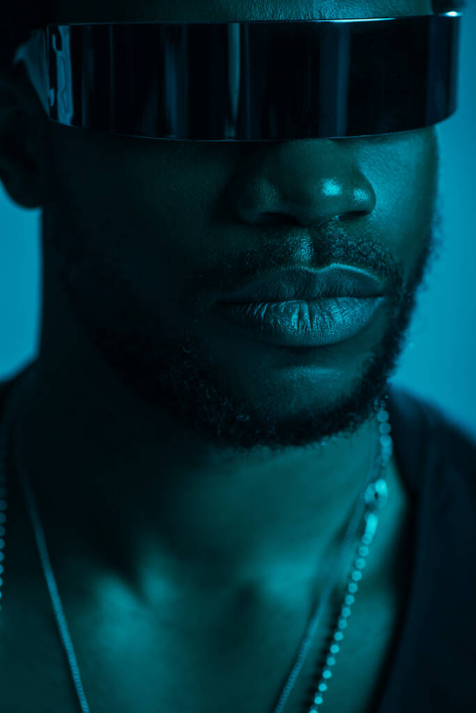 schöner afrikanisch-amerikanischer Mann mit futuristischer Sonnenbrille posiert isoliert auf blauem Grund in blauem Licht  - Foto, Bild