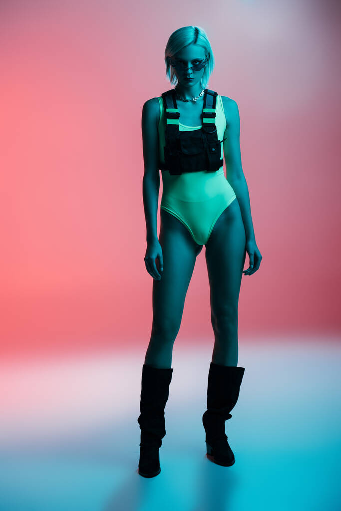 fashionable girl in futuristic bodysuit and fire-shaped sunglasses posing on pink in blue light  - Photo, Image