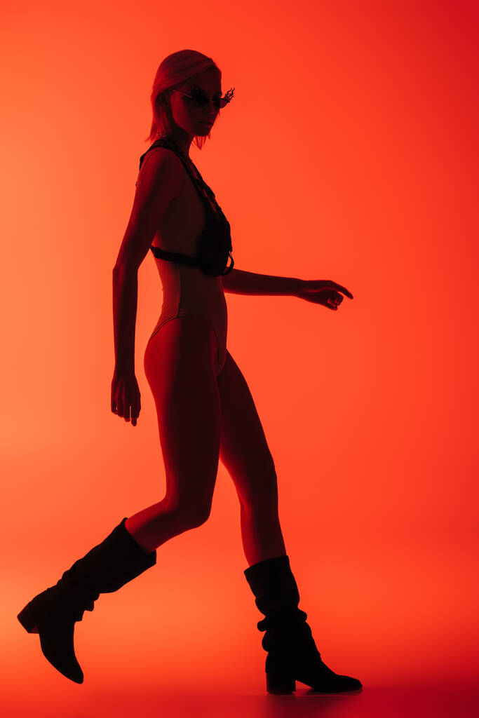 fashionable futuristic girl posing in bodysuit and fire-shaped sunglasses on red - Photo, Image