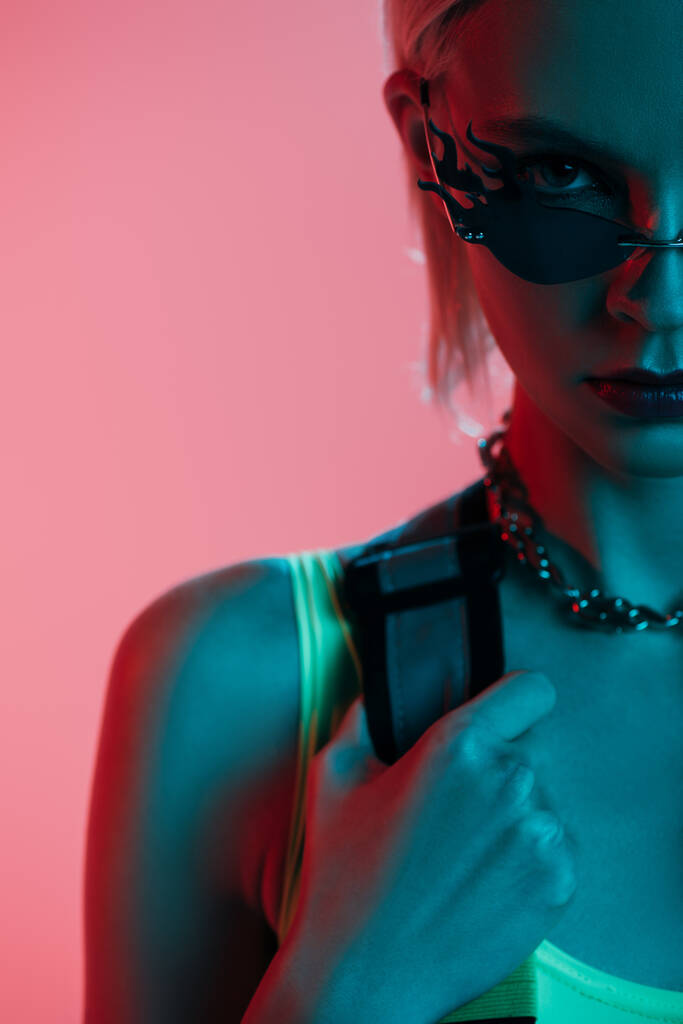 modelo elegante em collants futuristas e óculos de sol em forma de fogo posando isolado em rosa em luz azul
  - Foto, Imagem