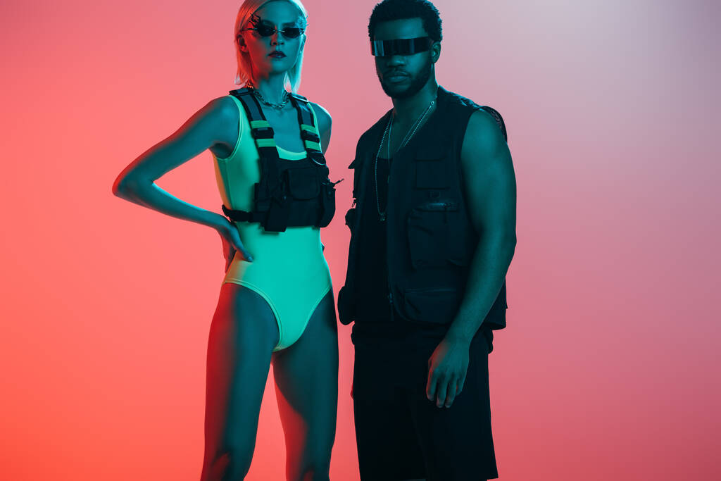 stylish multicultural couple posing in futuristic look and sunglasses on pink - Photo, Image