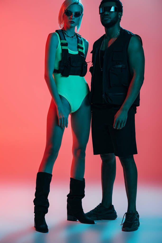 pareja interracial de moda posando en look futurista y gafas de sol en rosa
 - Foto, Imagen