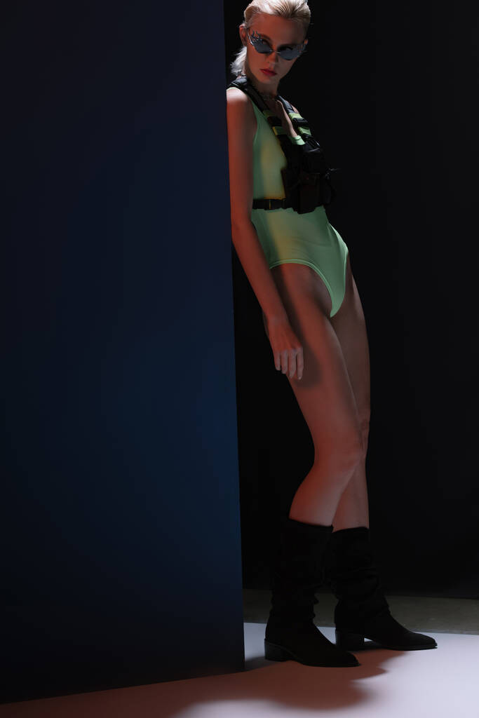 gyönyörű elegáns lány futurisztikus testöltöny és tűz alakú napszemüveg pózol a sötét  - Fotó, kép