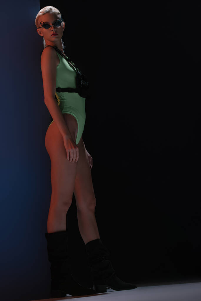 привлекательная модная девушка в футуристическом телесном костюме и солнцезащитных очках в форме огня, позирующих на темноте
  - Фото, изображение