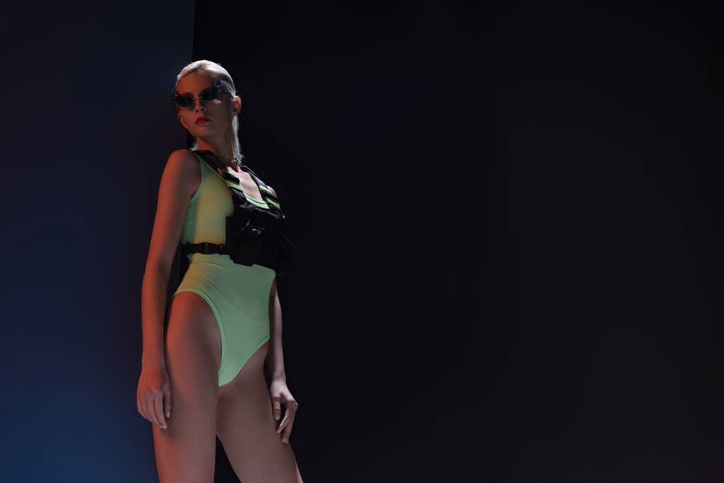 krásná módní žena v futuristickém obleku a ohnivé sluneční brýle pózující na tmavé  - Fotografie, Obrázek