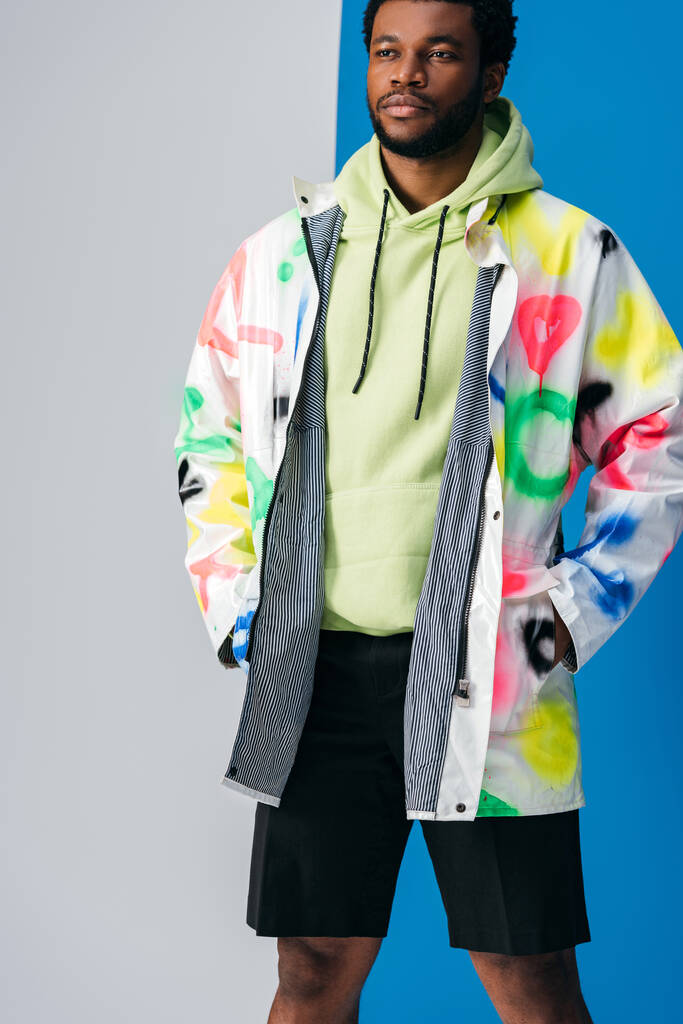 przystojny młody Afroamerykanin człowiek pozowanie w kolorowe futurystyczne ubrania na szary i niebieski - Zdjęcie, obraz