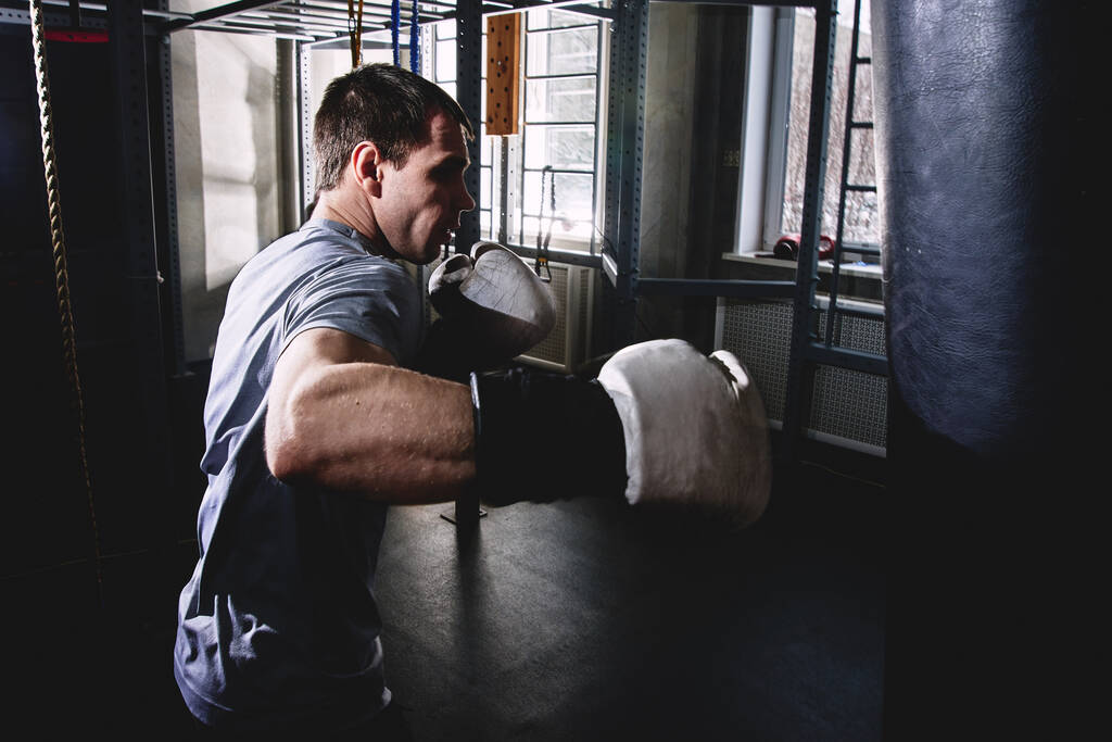 Homem musculoso boxeador profissional bate um saco de perfuração em luvas de boxe, treinando um atleta no ginásio, uma batalha agressiva em cores escuras
 - Foto, Imagem