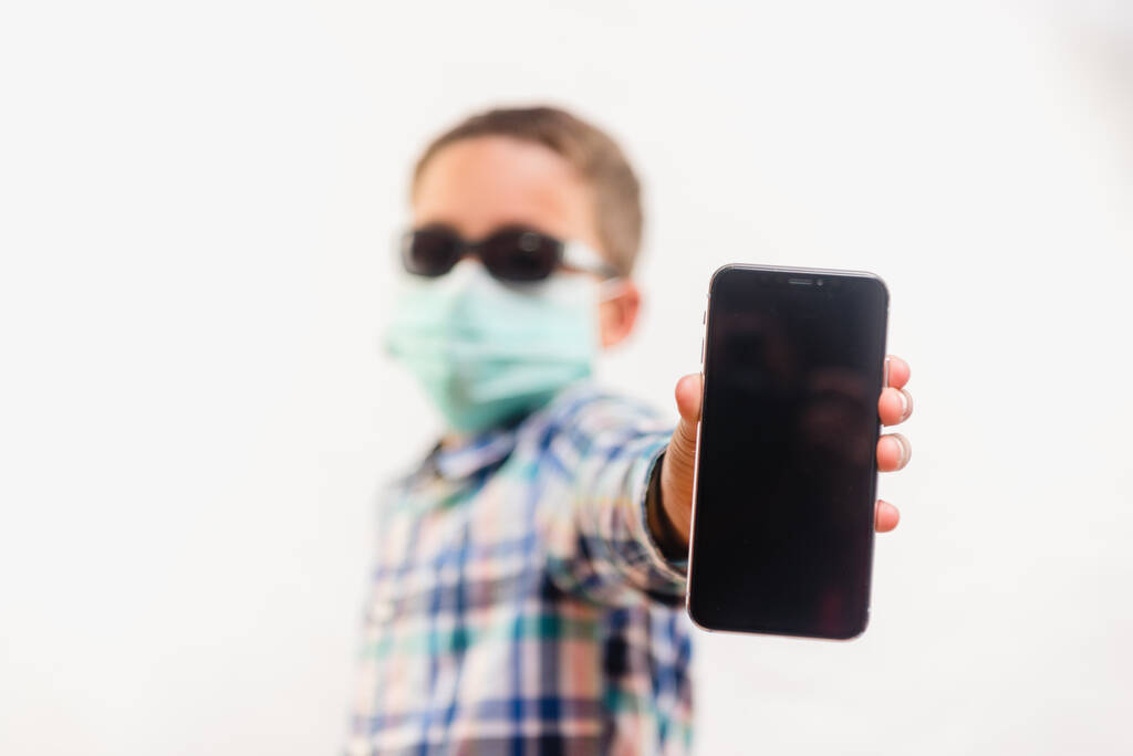Мальчик в одноразовой маске и солнцезащитных очках держит мобильный телефон на белом фоне
 - Фото, изображение