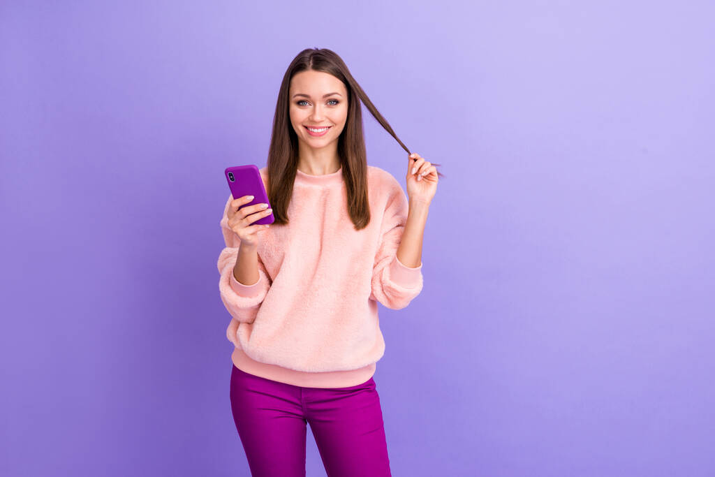 美しいブロガーの女性の携帯電話の手を持っているの写真長いカール着用カジュアル毛皮のふわふわ柔らかいプルオーバーパンツ孤立紫色の色の背景と遊ぶ友人をテキストメッセージ - 写真・画像