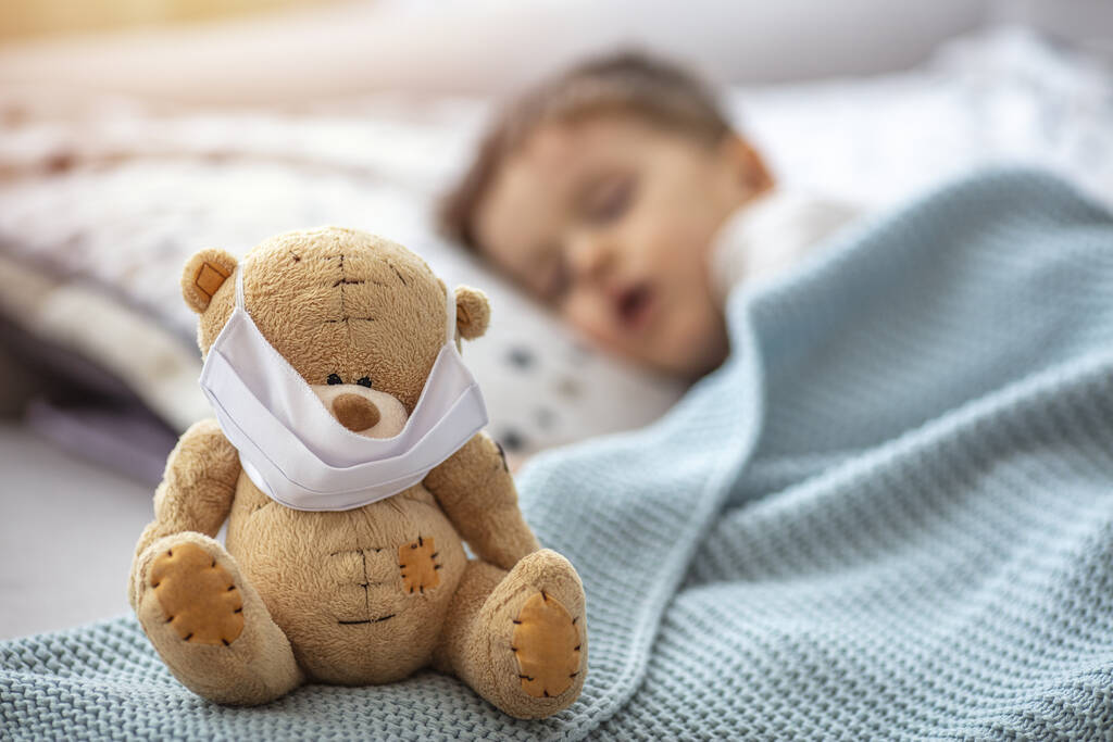 彼の病気のテディベアの医療マスクで、ベッドでの家庭の隔離での子供は、コロナウイルスCOVID-19とインフルエンザの流行の間のウイルスに対する保護のために、寝ています。子供と病気の概念 - 写真・画像