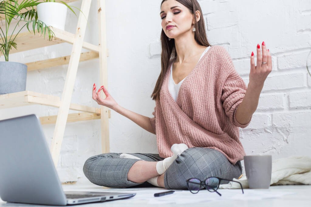 Młoda szczęśliwa kobieta siedzi na podłodze w mieszkaniu do jogi w jasnym mieszkaniu i pracuje w laptopie, freelancerka medytuje, podczas gdy praca stresuje się - Zdjęcie, obraz